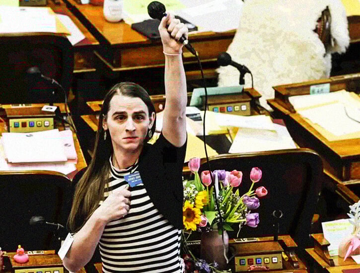 A woman on the floor of the Montana House holds up a microphone, her right hand clutched against her chest, in protest.